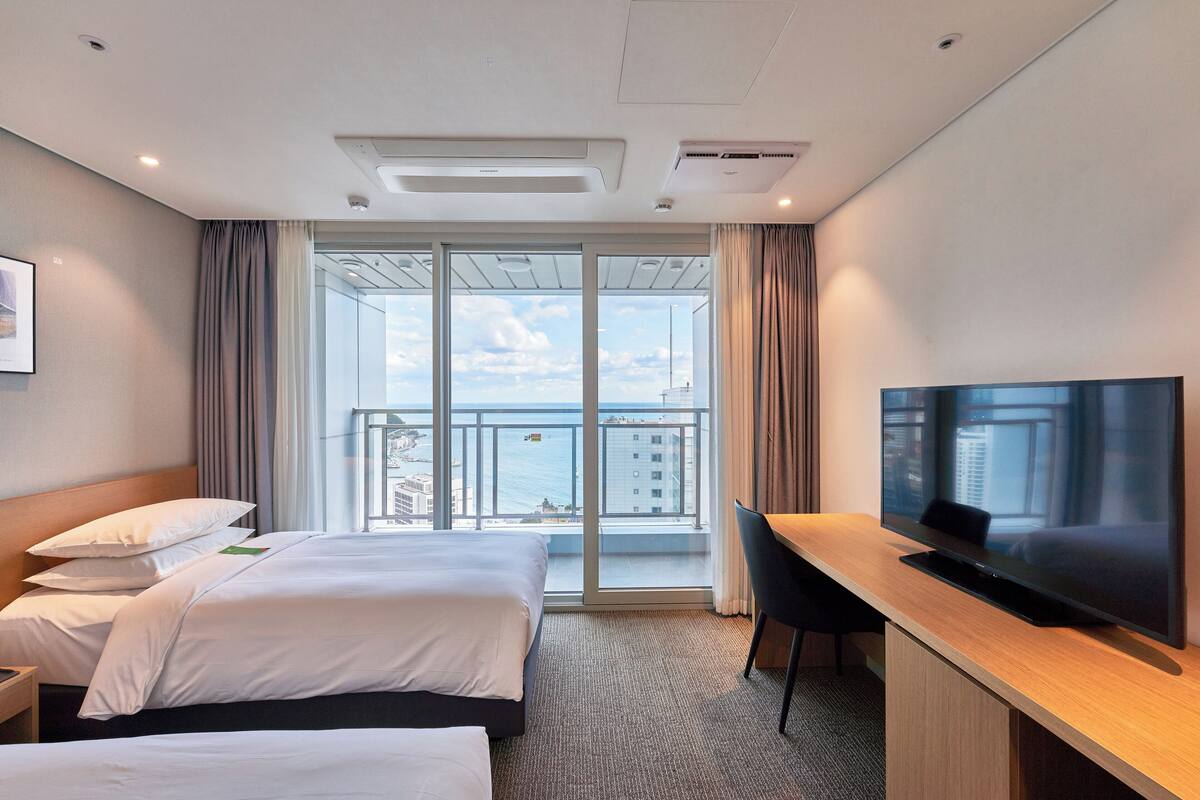 A hotel room with a large bed, a desk with a TV, and a balcony with a view of the city.
