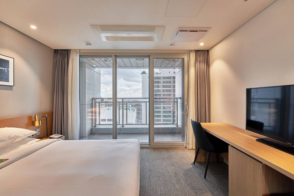 A hotel room with a bed, a desk with a chair, a television, and a large window with a view of buildings.
