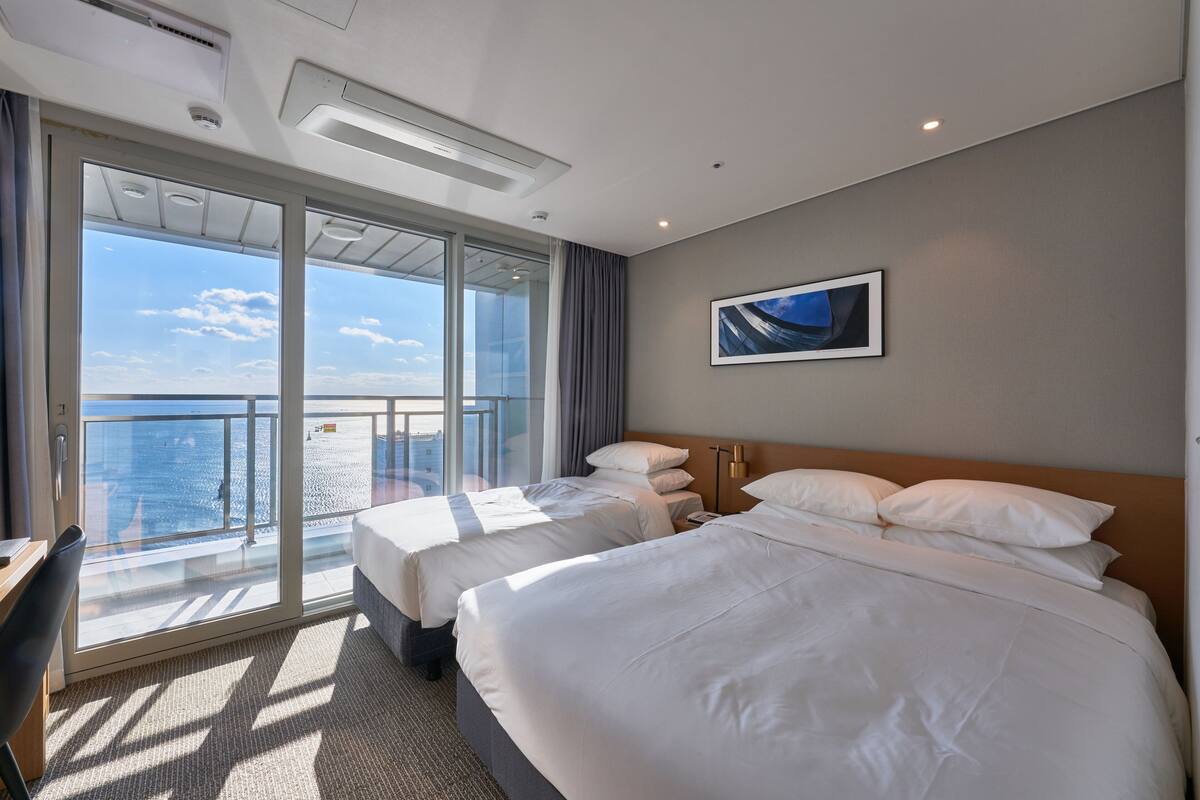 A hotel room with two beds, a large window with a view of the sea, and a painting on the wall.