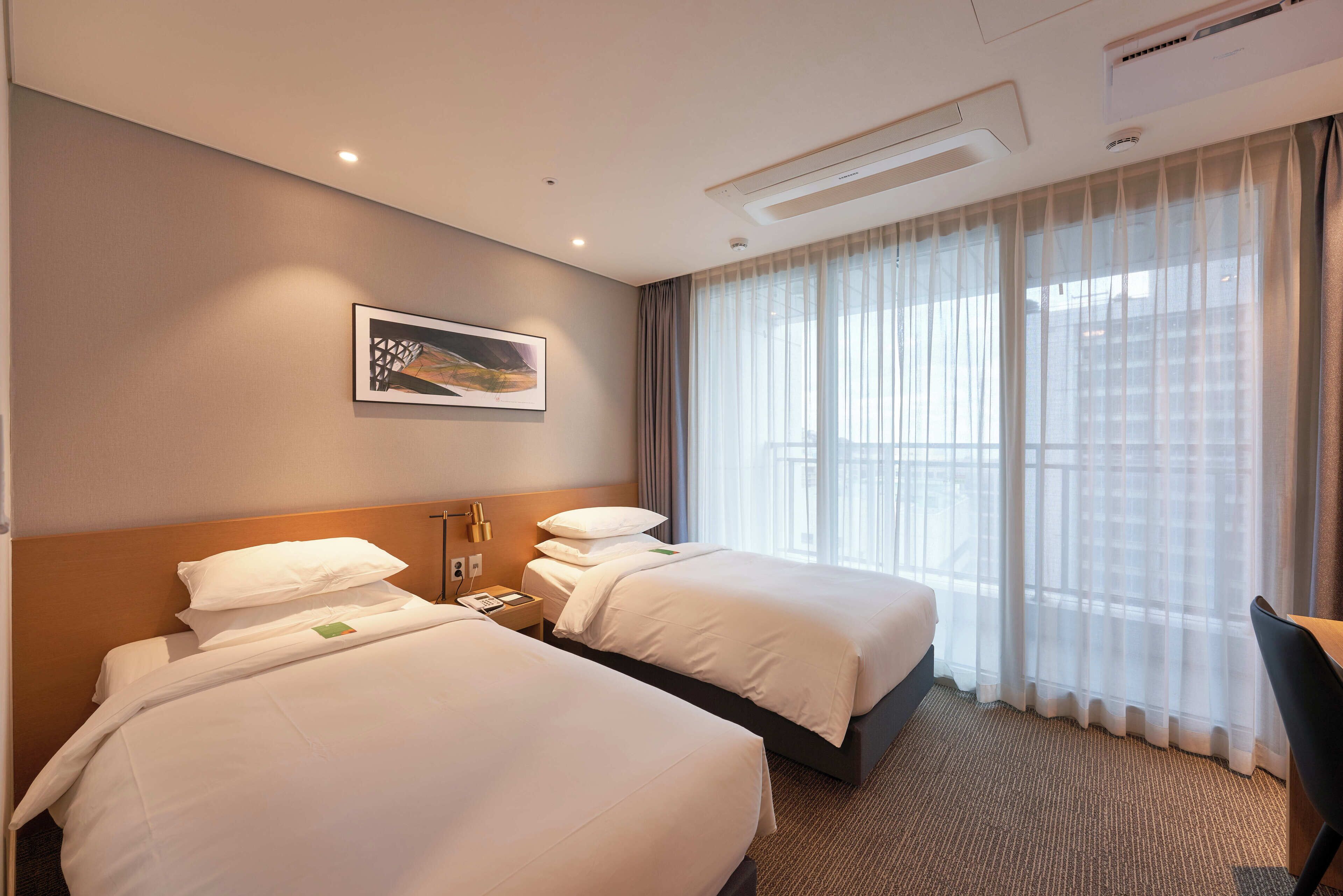 Quarto de hotel com duas camas, uma janela grande com cortinas translúcidas, um quadro na parede e ar condicionado no teto.