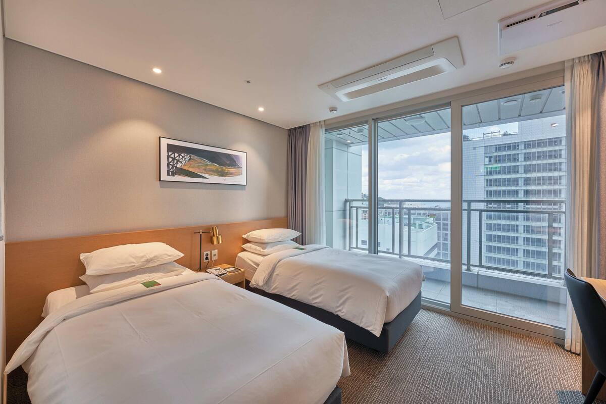 A hotel room with two beds, a large window with a city view, and a framed picture on the wall.