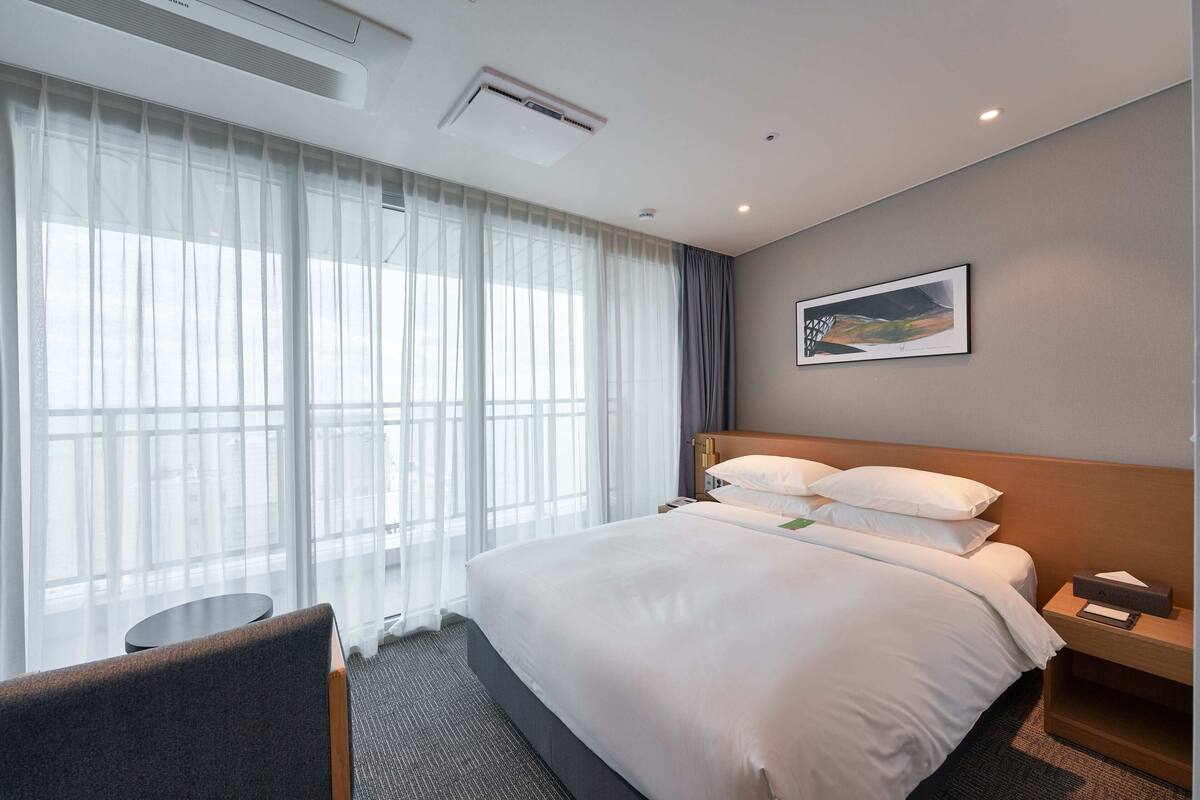 A hotel room with a large bed, a bedside table, a chair, and a window with sheer curtains.