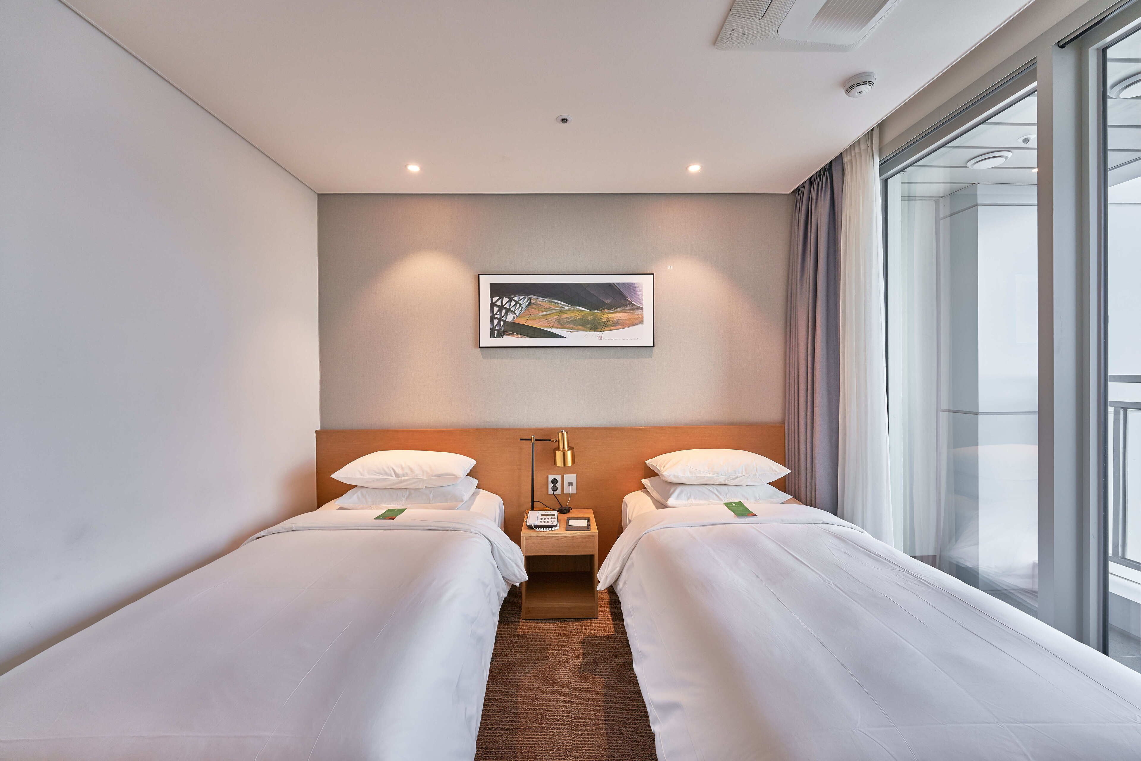 A hotel room with two single beds, a nightstand with a lamp, a painting on the wall, and a large window with curtains.