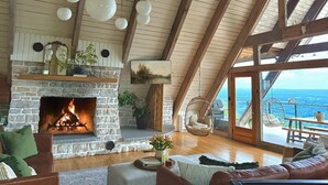 A cozy living room with a stone fireplace, wooden beams, hanging spherical lights, and a large window overlooking a scenic view. - A-frame W/ Hot-tub, Mountain View & tennis court (Newport)