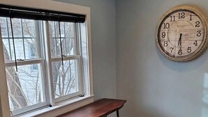 Una habitación con una mesa de madera y tres taburetes, una ventana con persianas, y un reloj de pared.