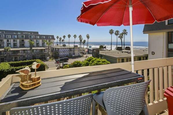 A balcony with a red umbrella, a table, and chairs overlooking a beach with palm trees and a pier.
