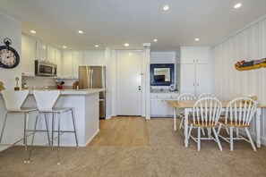 A compact kitchen with a microwave, refrigerator, and a sink. There are two white bar stools and a wooden dining table with four chairs. A mirror hangs above the sink, and a clock is on the wall.