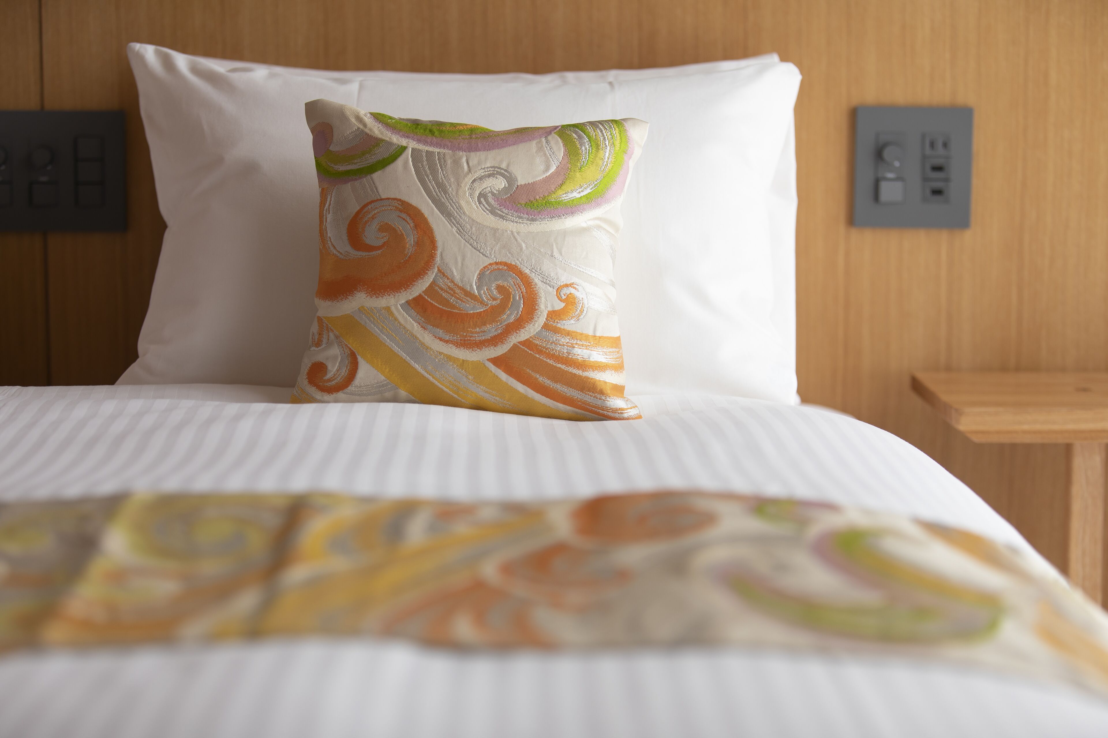 Ein ordentlich gemachtes Bett mit weißer Streifenbettdecke und farbigem Kopfkissen.