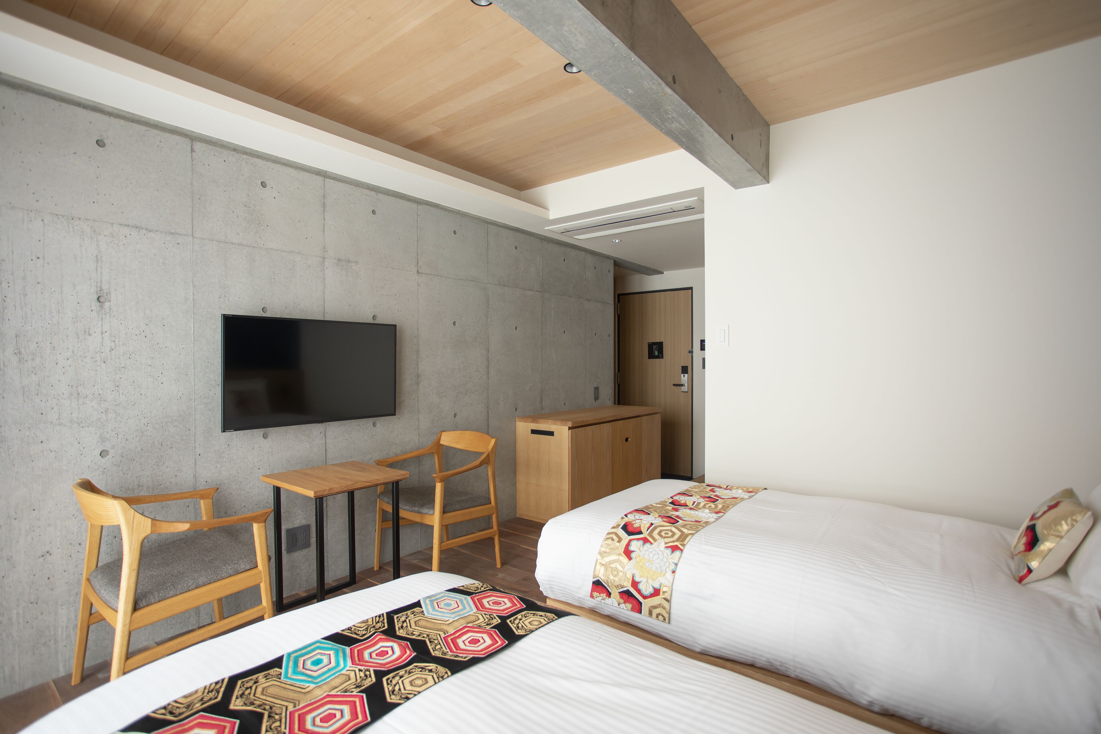 A modern hotel room with two beds, a flat-screen TV, wooden furniture, and a concrete accent wall.