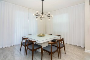Un coin repas avec une table en verre, six chaises en bois, un lustre et des rideaux blancs.