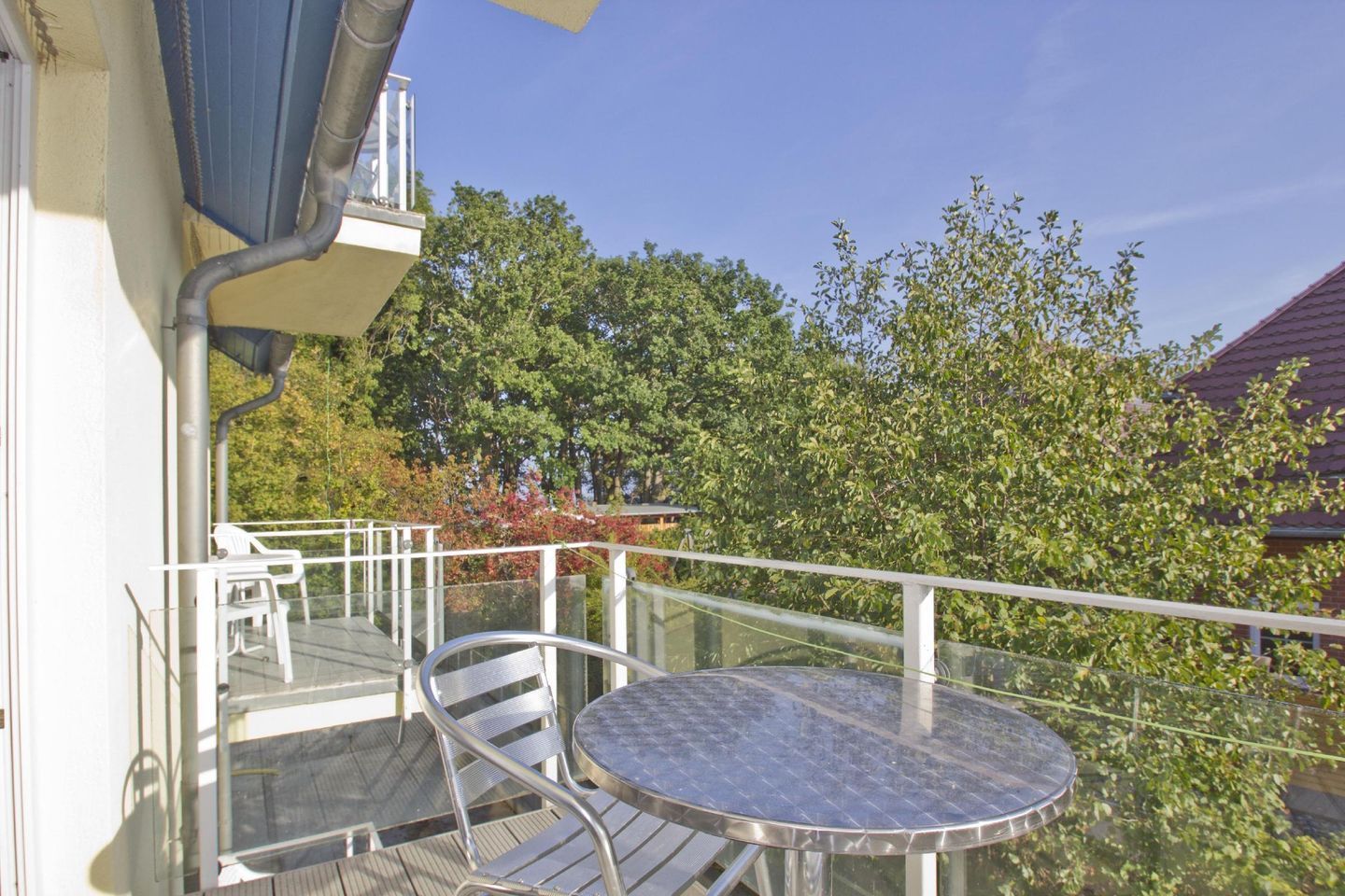 Un balcon avec une table et des chaises en verre, donnant sur un jardin avec des arbres et une maison en arrière-plan.