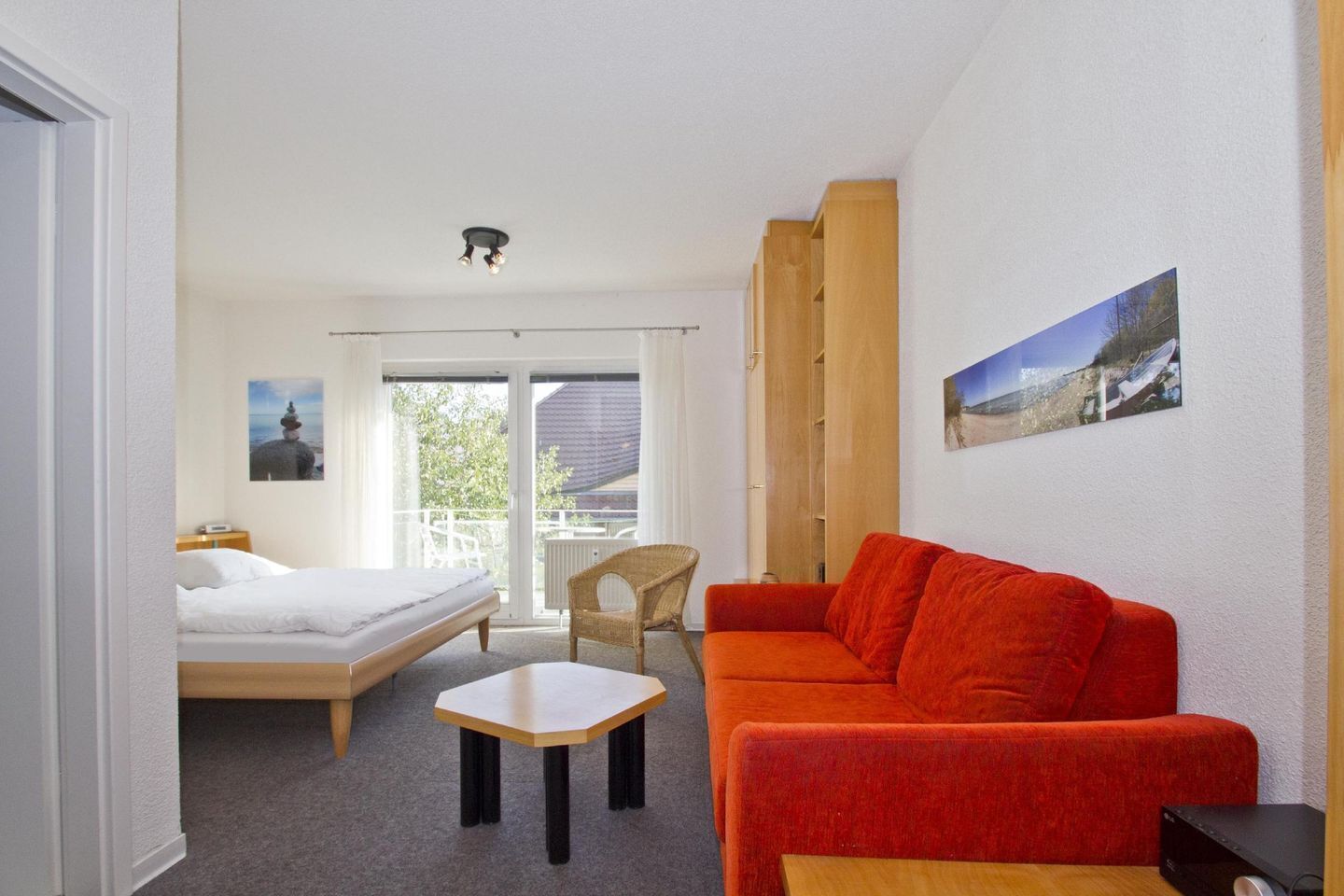 Une chambre d’hôtel avec un lit, un canapé rouge, une petite table et une chaise. Il y a un balcon avec vue sur l’extérieur.