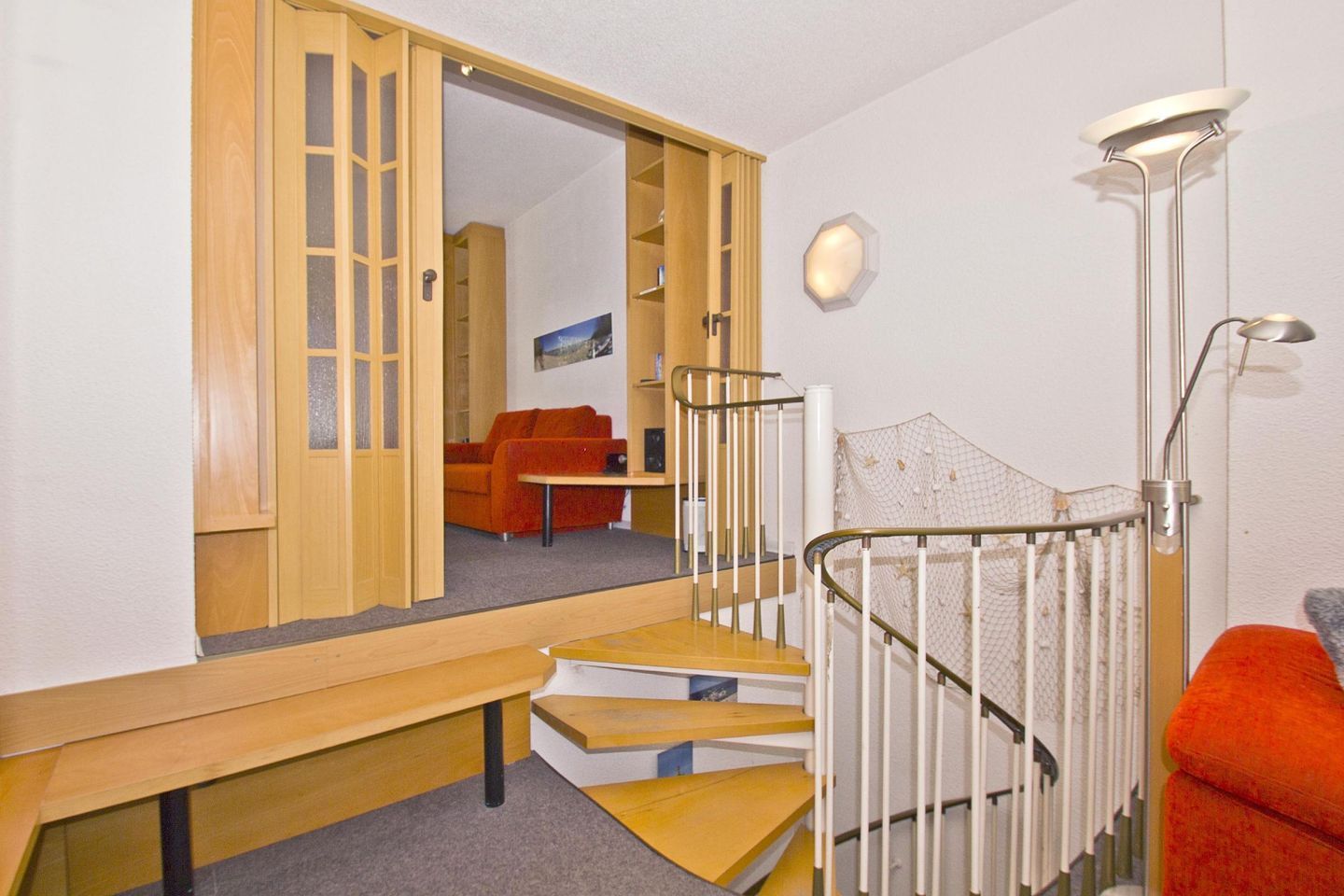 Een kamer met een houten bank, een trap met een witte leuning en een rode bankstel op de achtergrond.