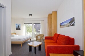 A hotel room with a bed, a red sofa, a small table, and a chair. There is a balcony with a view of the outside.