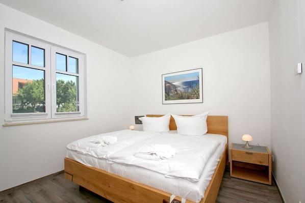 A bedroom with a wooden bed, white bedding, a nightstand with a lamp, a window with a view of trees and buildings, and a framed picture on the wall.