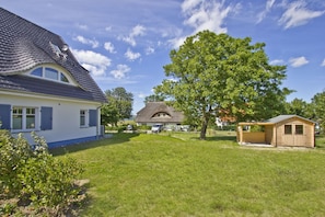Une maison à l’extérieur bleu, une construction à toit de chaume en arrière-plan, un abri en bois et un grand arbre dans le jardin.