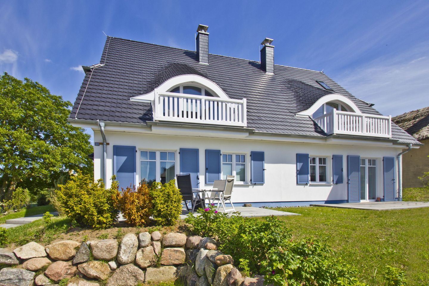 A two-story house with a balcony, a stone wall in the foreground, and a well-maintained garden.