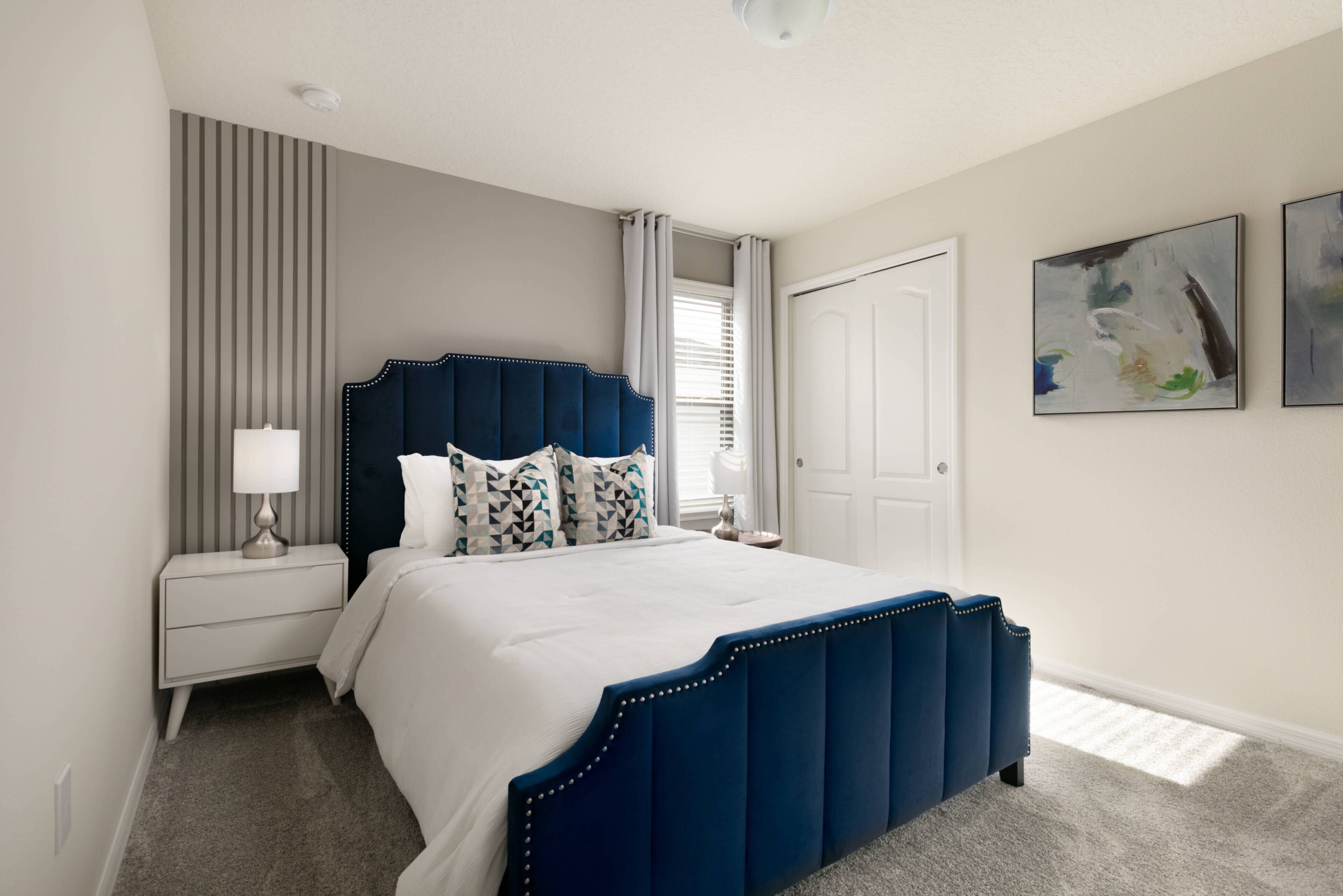A bedroom with a large bed, a nightstand, a lamp, a window with curtains, and two abstract paintings on the wall.