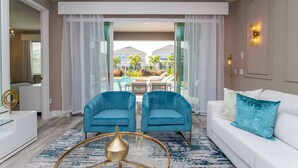 A modern living room with a white sofa, two blue armchairs, and a glass coffee table. A large window offers a view of a pool and buildings. - Family Escape With Private Pool & Themed Rooms (Kissimmee)
