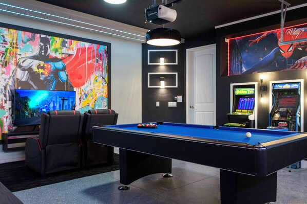 A pool table with a blue felt surface, two black chairs, a projector mounted on the ceiling, and arcade machines in the background. - Family Escape With Private Pool & Themed Rooms (Kissimmee)