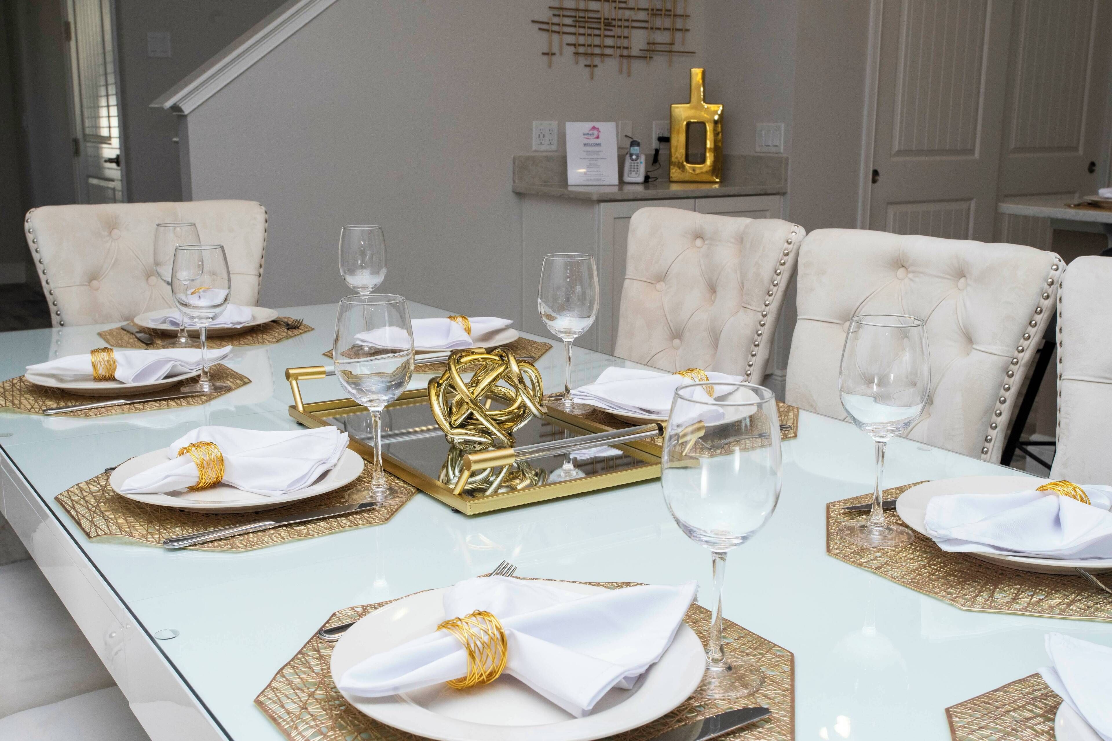 A dining table set with white plates, gold napkin rings, and clear wine glasses, surrounded by tufted chairs.