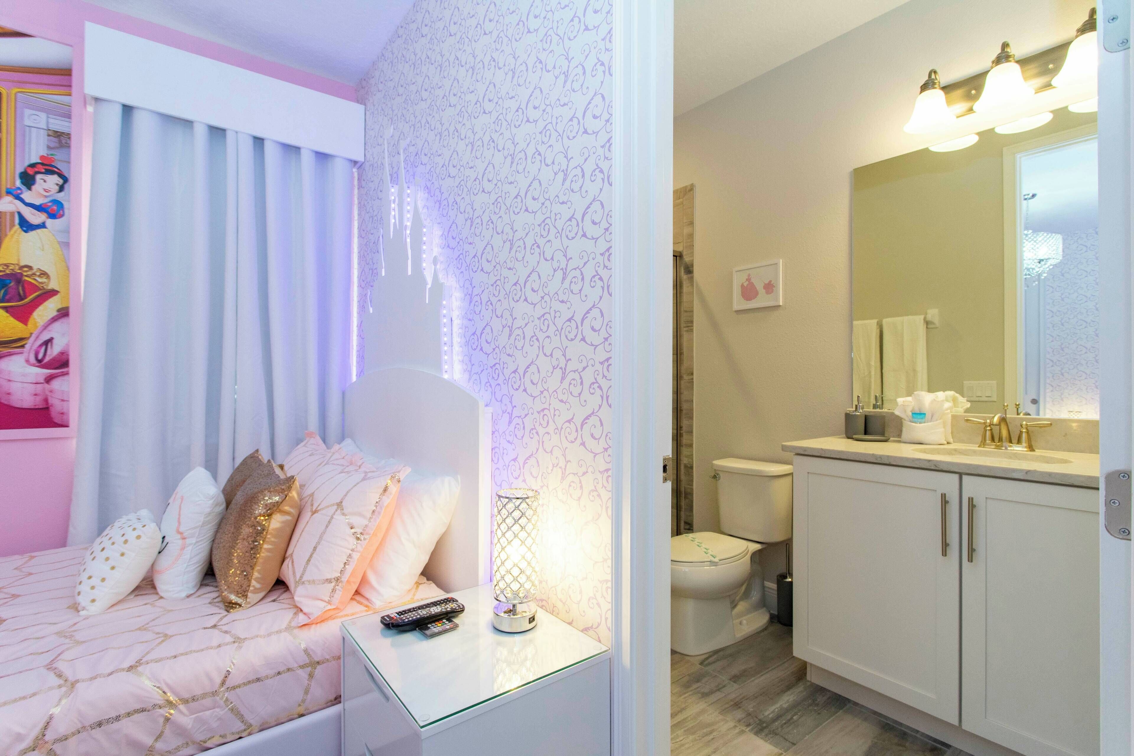 Et soveværelse med seng, sengebord og et natbord med lampe. Der er et badeværelse med toilet, håndvask og spejl.
