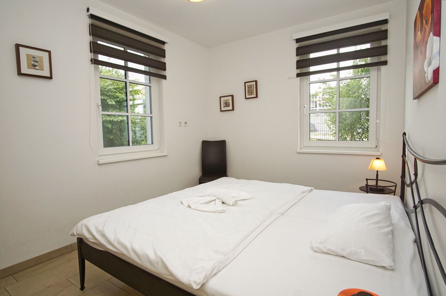 Un dormitorio con cama, dos ventanas con persianas, una silla, una mesita de noche con lámpara y cuadros en la pared.