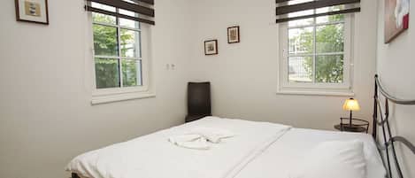 Un dormitorio con cama, dos ventanas con persianas, una silla, una mesita de noche con lámpara y cuadros en la pared.