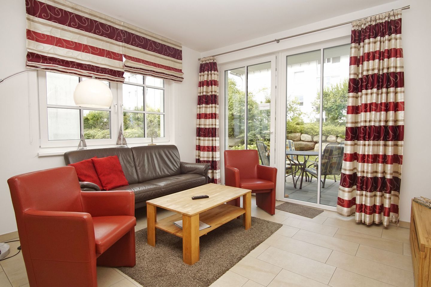 Ein Wohnzimmer mit braunem Ledersofa, roten Sesseln, einem Holztisch und einem großen Fenster mit Vorhängen.