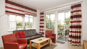 A living room with a brown leather sofa, red chairs, a wooden coffee table, and a large window with curtains. - Terrace, free use of the AHOI adventure pool and sauna - Villa Antje FeWo 04 - (Göhren)