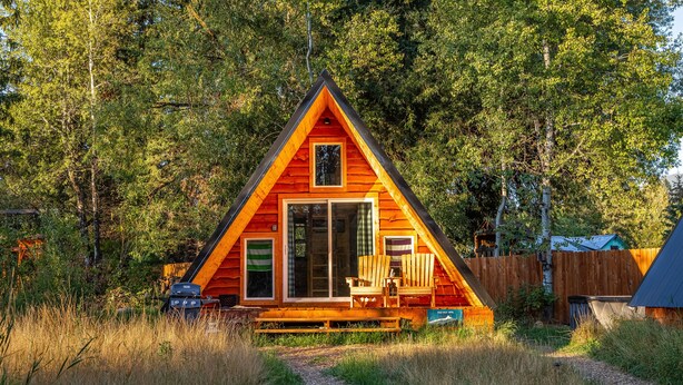 Una cabaña de madera triangular con terraza, rodeada de árboles altos.