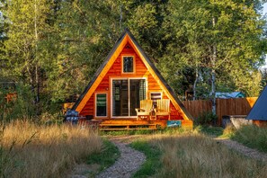 A triangular wooden cabin with a deck, surrounded by tall trees.