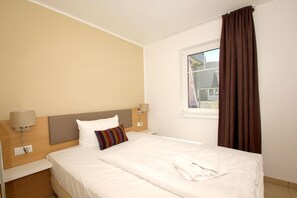 A hotel room with a bed, a window with curtains, a bedside lamp, and a wall-mounted control panel.