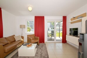 Un salon moderne avec un canapé en cuir marron, une table basse blanche, un rideau rouge, un téléviseur et une porte-fenêtre coulissante donnant sur l’extérieur.