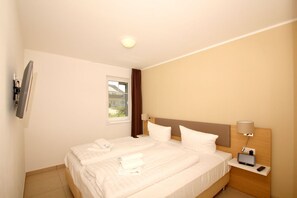 A hotel room with a bed, a TV mounted on the wall, a window with curtains, and a bedside table with a lamp and a remote control. - Beachfront vacation rental with a terrace, access to the pool and sauna - Rex Rugia H-1010 - (Göhren)