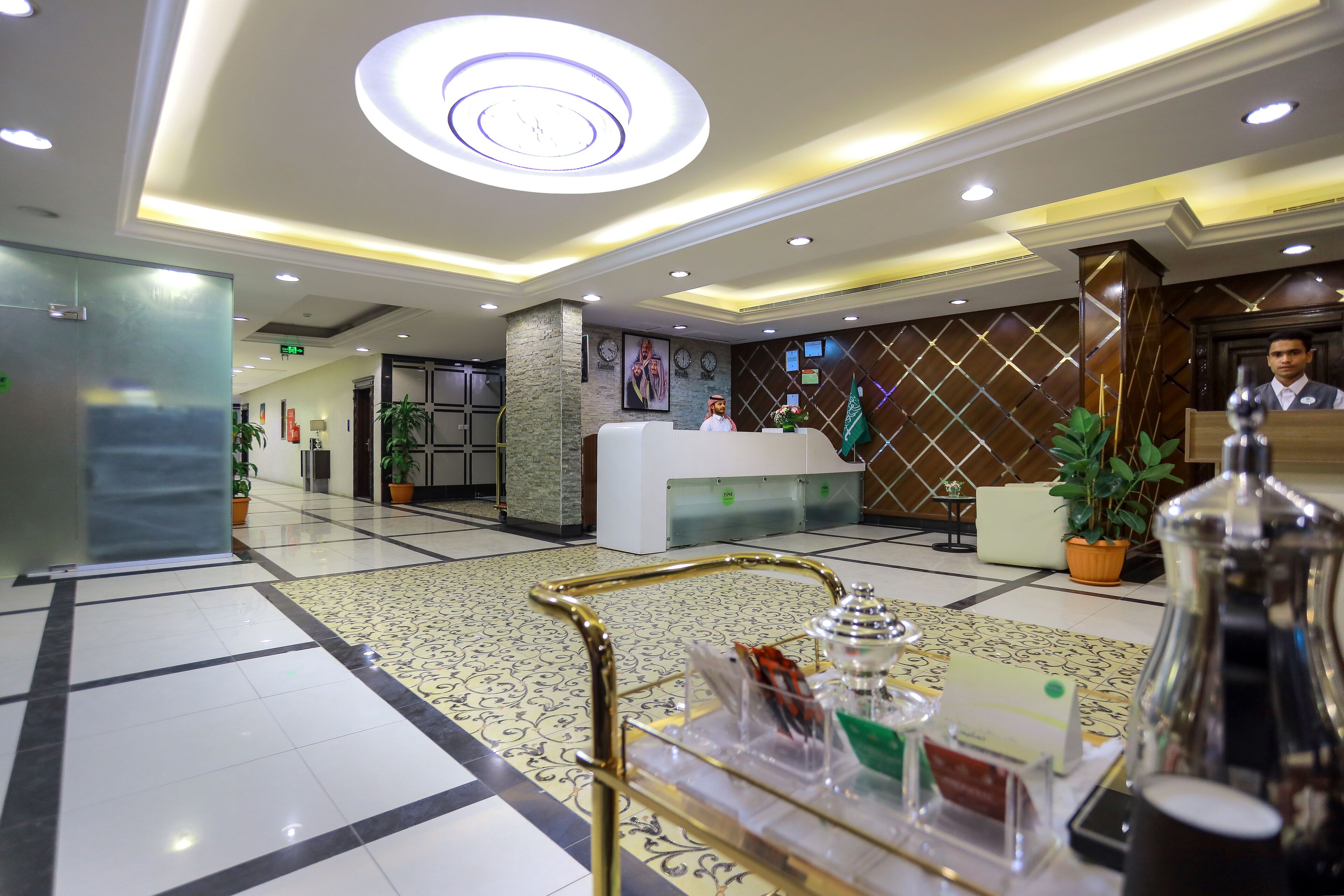Un vestíbulo de hotel con mostrador de recepción, una mampara de cristal y una lámpara decorativa en el techo.
