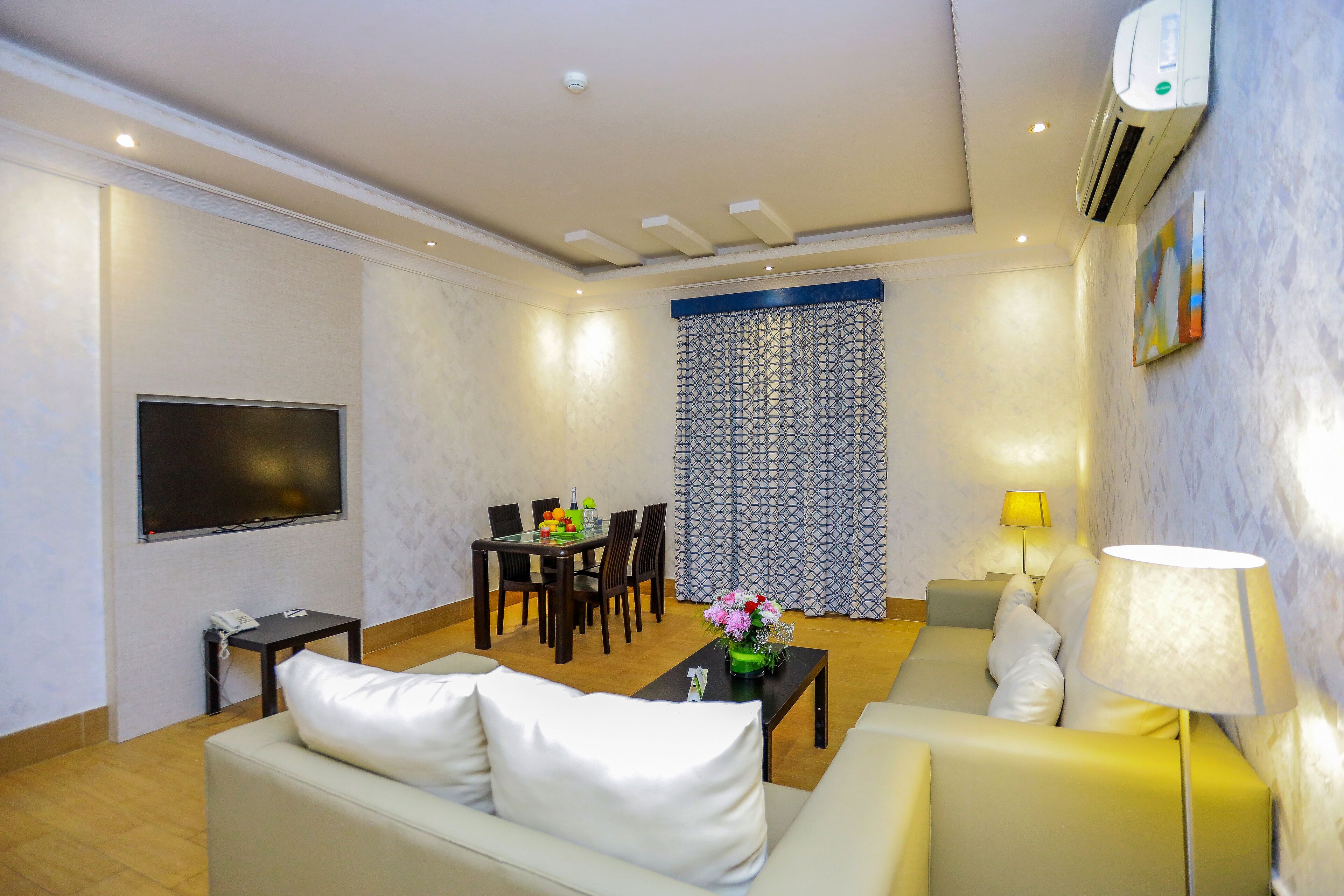 A modern living room with a sofa, a coffee table with flowers, a dining area with chairs, a TV, and an air conditioning unit.