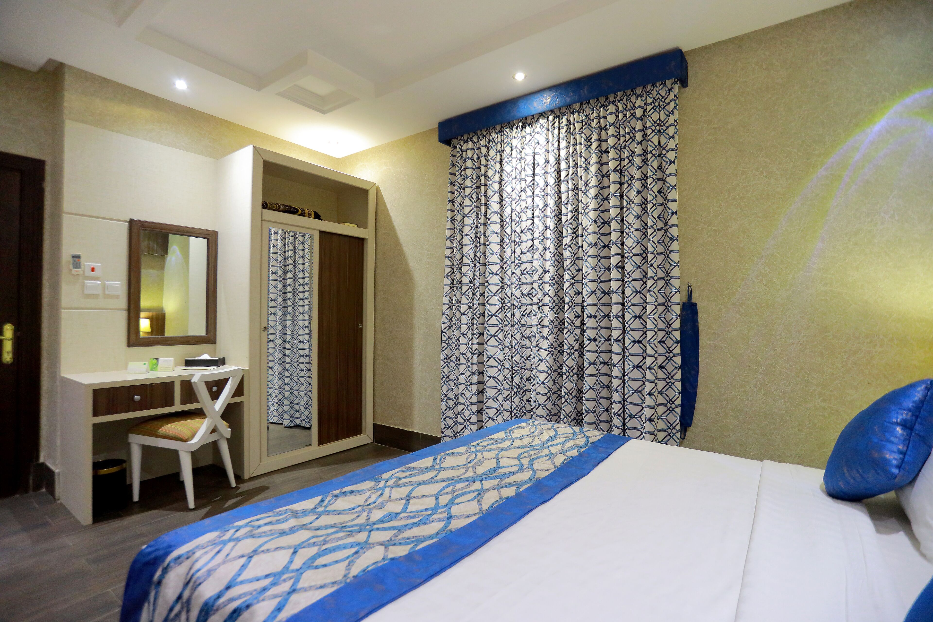 A hotel room with a bed, a desk, a chair, a wardrobe, and a curtain with a geometric pattern.