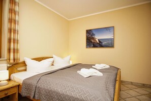 Habitación de hotel con cama, mesita de noche, lámpara, ventana con cortinas y un cuadro de un paisaje costero.