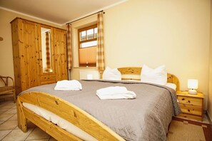 A wooden bed with a grey blanket, two white towels, a wooden nightstand with a lamp, and a wooden wardrobe.