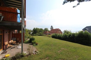 A residential property with a two-story building, a covered patio with outdoor furniture, a well-maintained lawn, and a view of a larger house across a grassy area.
