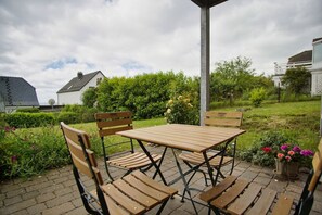 An outdoor patio with a wooden table and chairs, surrounded by greenery and a hedge.