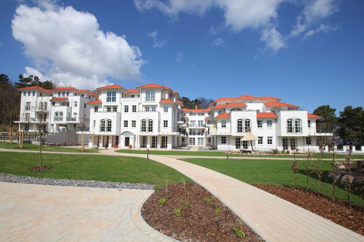 A large, multi-story building with a red-tiled roof, white exterior walls, and a paved walkway leading to the entrance.