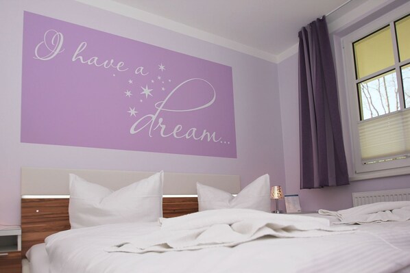 A bedroom with a bed, white bedding, a headboard, a nightstand, a wall with a purple sign, a window with curtains, and a bedside lamp.