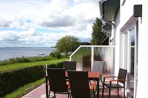 Une terrasse extérieure avec une table et des chaises, donnant sur une étendue d’eau et une zone herbeuse.