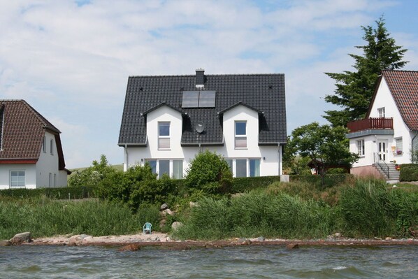 Une maison à deux étages, avec un toit recouvert de tuiles sombres, des murs blancs et un petit balcon au deuxième étage.