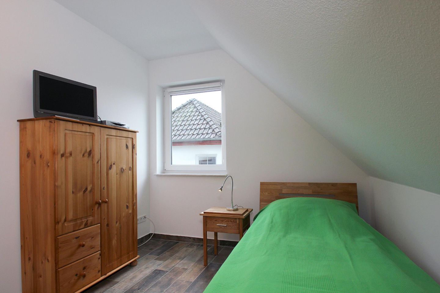 A small, tidy room with a wooden wardrobe, a single bed with a green blanket, a nightstand with a lamp, and a window with a view of a house.