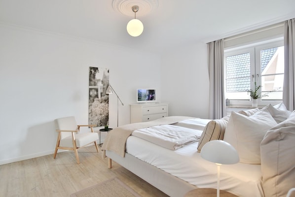 A modern bedroom with a bed, bedside table, chair, and a large window with curtains.