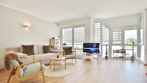 A modern living room with a sofa, two armchairs, a coffee table, and a TV on a stand. Large windows provide natural light and views of the outside. - Gorch-Fock-Str 16 Apartment 6 - Gorch-Fock-Str 16 (Timmendorfer Strand)