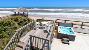 Une terrasse en bois avec un jacuzzi, des chaises et un belvédère offrant une vue sur la plage.